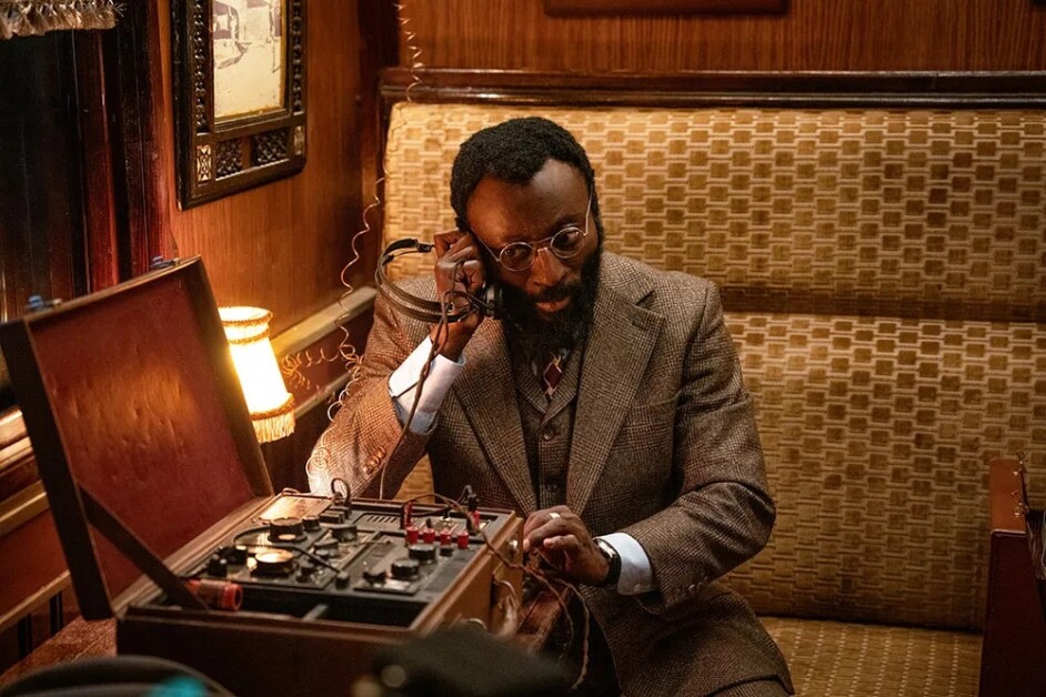 A man with glasses and a beard uses vintage radio equipment in a wooden booth, dressed in a brown suit.