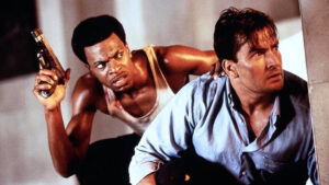 ‘Money Talks’ Blu-Ray Review – Chris Tucker Works Overtime For Action-Comedy Outing Two men crouch behind a wall, one holding a handgun and looking intense with a tank top, and the other appearing wary in a button-up shirt.