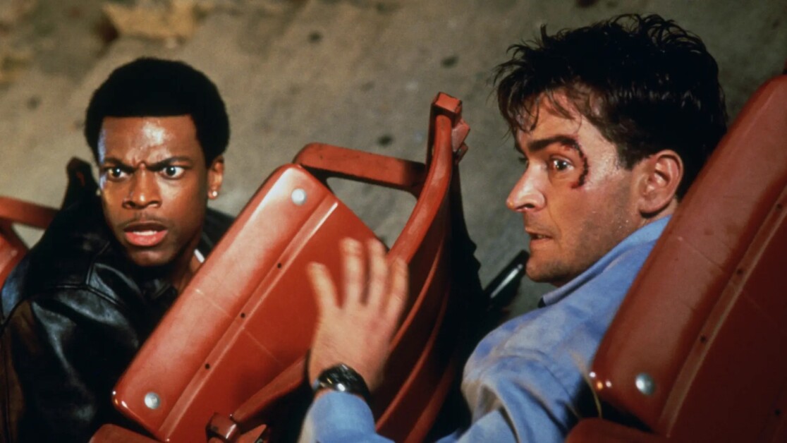 Two men sit in red seats, looking startled. One man on the left wears a black jacket, while the other on the right has a bruised face and wears a blue shirt.