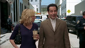 ‘Monk’: Seasons 5-6 (Individual) Blu-Ray Review – More Small Screen Mysteries With Our Favorite Detective A man in a beige suit and a woman in a purple top walking and talking on a city street, with a police officer in the background.