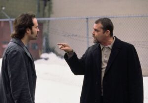 Two men are standing outside near a chain-link fence. One man with a beard and coat is pointing aggressively at the other man with longer hair and a goatee. Snow is visible on the ground.