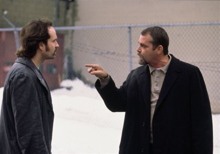 Two men are standing outside near a chain-link fence. One man with a beard and coat is pointing aggressively at the other man with longer hair and a goatee. Snow is visible on the ground.