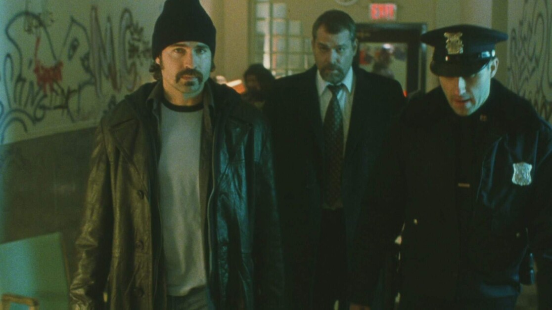Three men walk down a graffiti-covered hallway; one wears a beanie and leather jacket, another a suit and tie, and the third a police uniform.