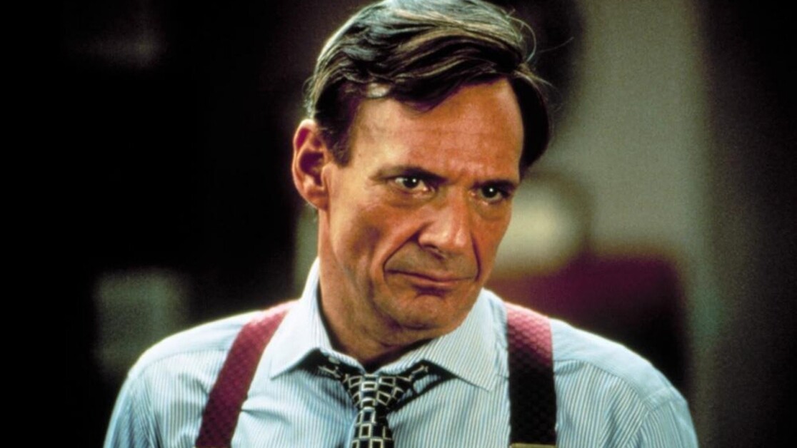 A man with short hair, wearing a striped shirt, red suspenders, and a loosely tied tie, looks with a serious expression.
