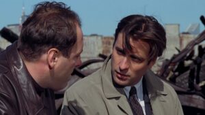 ‘Night Falls On Manhattan’ Arrow Video Blu-Ray Review – Blistering Crime Drama Explores The Thin Line Of Morality Two men engaged in a serious conversation outdoors; one is wearing a leather jacket, and the other a tan trench coat with a tie. Rusted metal debris is visible in the background.