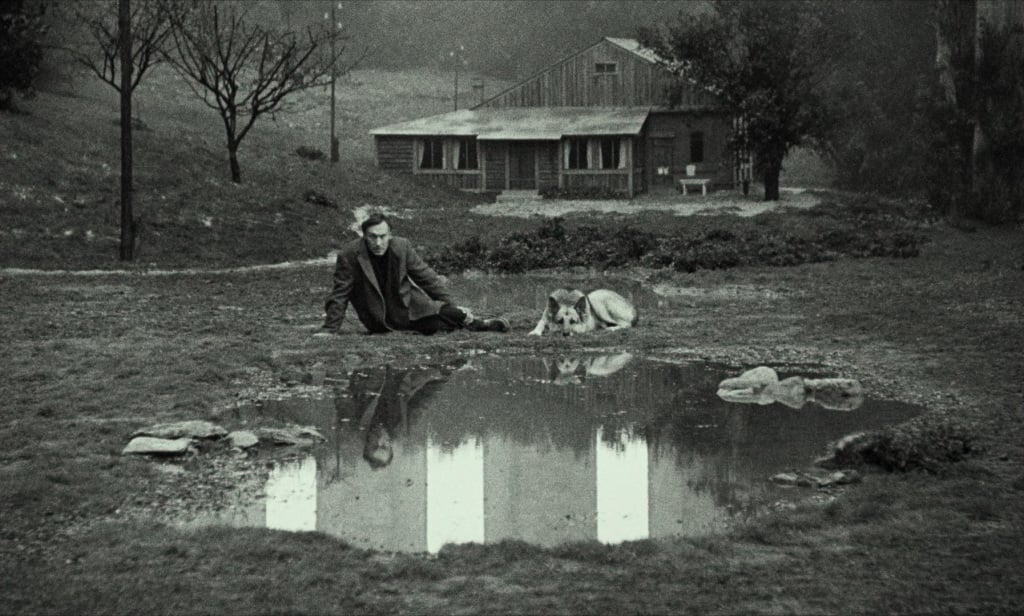 A black and white photo of a man sitting beside a reflective pond with a dog, looking at the water, with a wooden house and trees in the background.