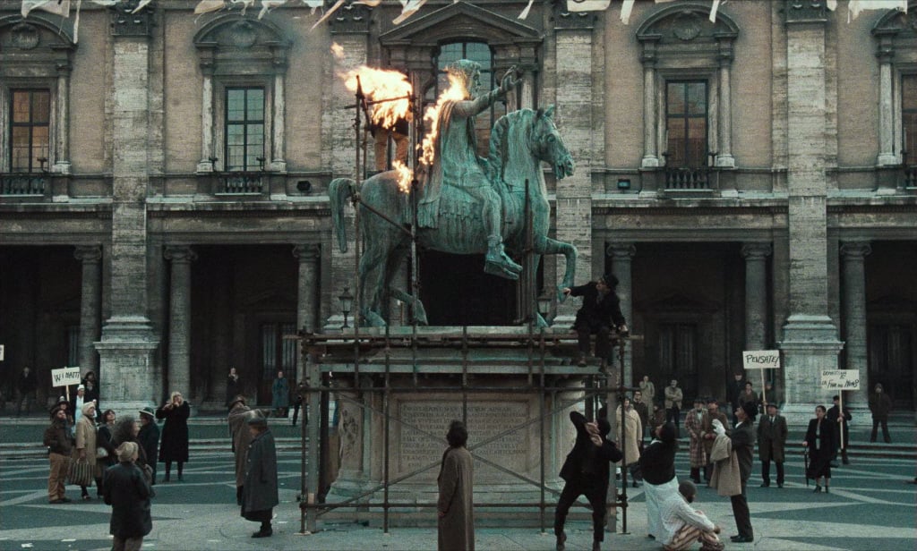 A bronze equestrian statue is set ablaze at night, surrounded by a crowd of onlookers in a large square.