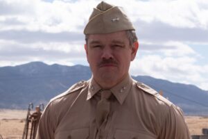 A man in a khaki military uniform and hat stands outdoors with mountains and a cloudy sky in the background.