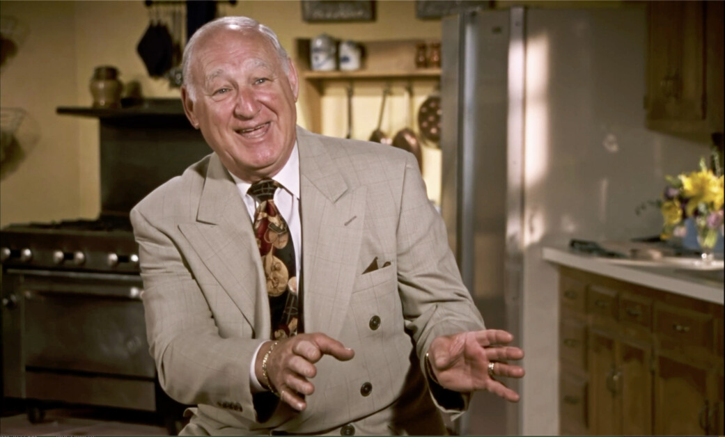 An elderly man in a beige suit and patterned tie is smiling and gesturing with both hands while seated in a kitchen setting.