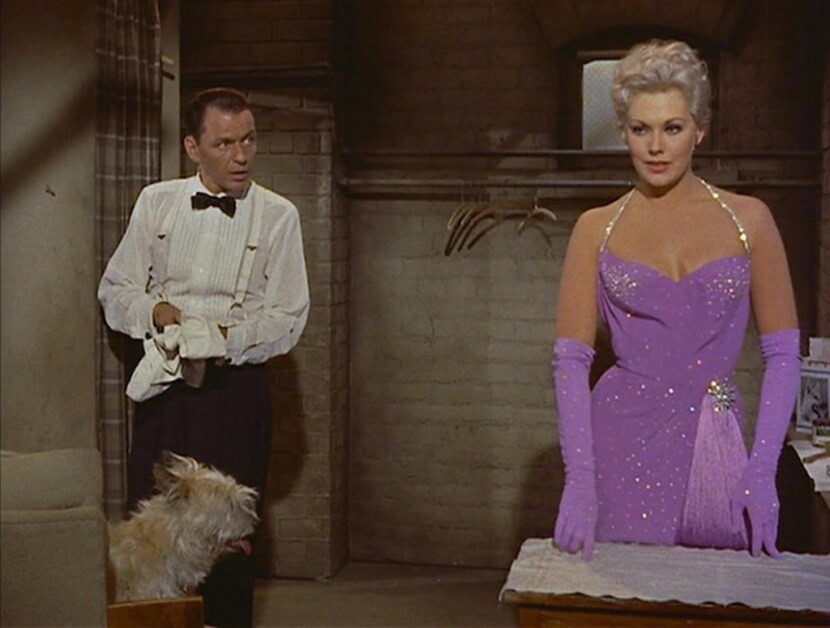 A man dressed in a white shirt and black bow tie stands next to a small dog, looking at a woman in a purple dress with matching gloves. The scene appears to be set in an indoor, brick-walled environment.