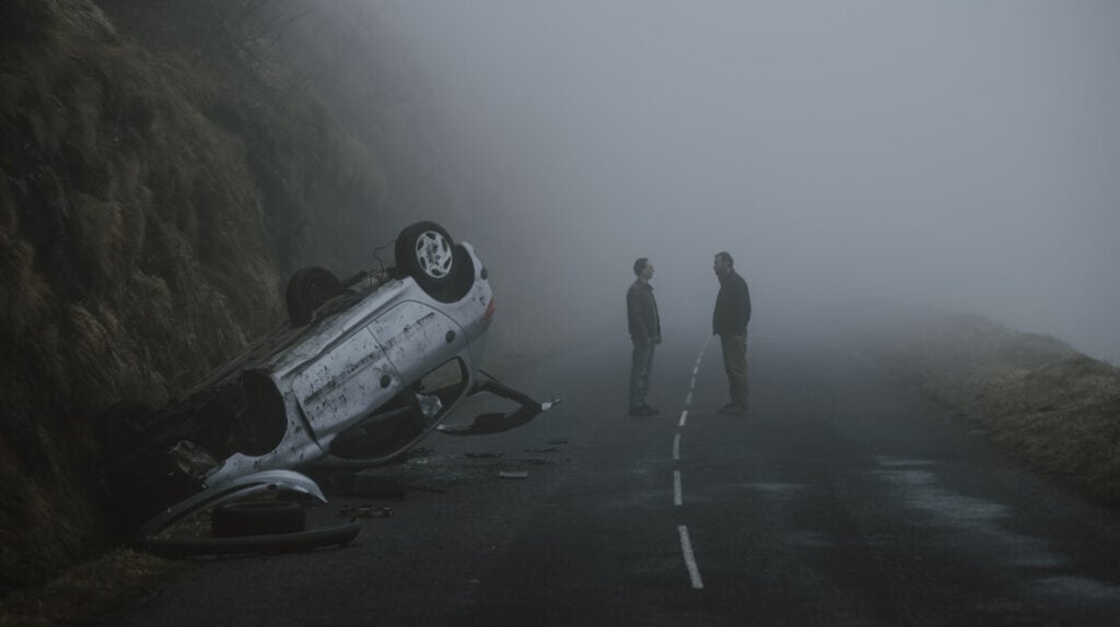 Two people stand facing each other on a foggy road next to an overturned car that has gone off an embankment. The scene appears tense and isolated.