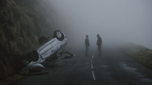 Two people stand facing each other on a foggy road next to an overturned car that has gone off an embankment. The scene appears tense and isolated.