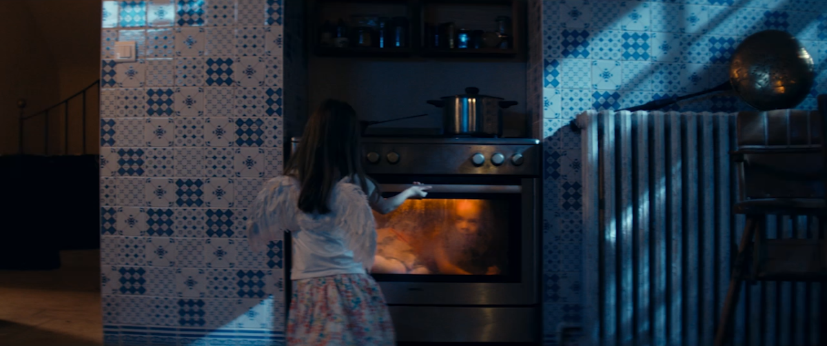 A young girl stands in a dimly lit kitchen, looking into an oven with illuminated contents. The walls feature a blue and white geometric tile pattern. A pot sits on the stove above.