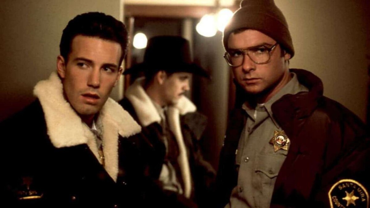 Two men in a hallway, one in a fur coat and the other in a beanie, both looking serious, with a third person slightly visible in the background.