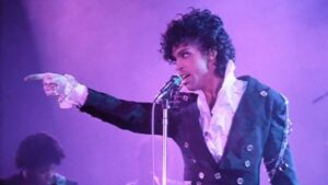 A man in a ruffled shirt and blazer sings into a microphone while pointing. The background is lit in pink and purple hues.