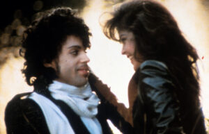 Two people stand close together, smiling at each other. The background features a bright, indistinct light that gives the scene a warm glow.