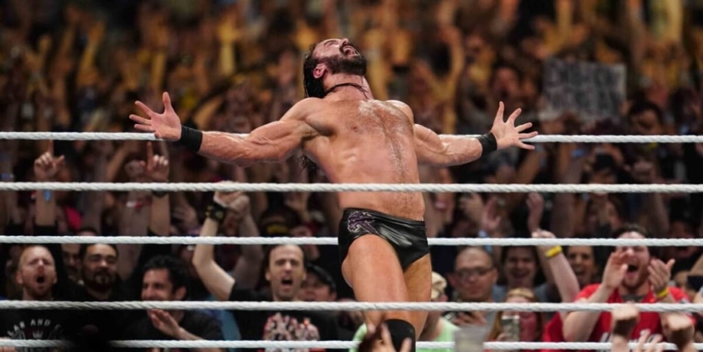 A wrestler celebrates victoriously in the ring, arms spread wide, with a cheering crowd in the background.