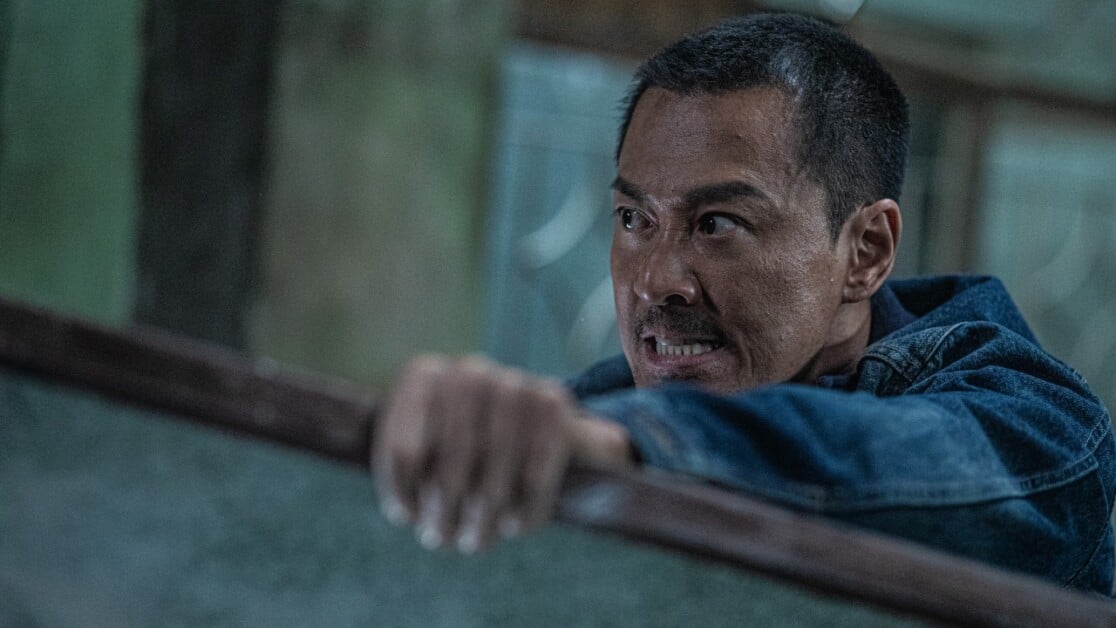 A man in a denim jacket is gripping a piece of wood and displays a determined, intense expression in a dimly lit environment.