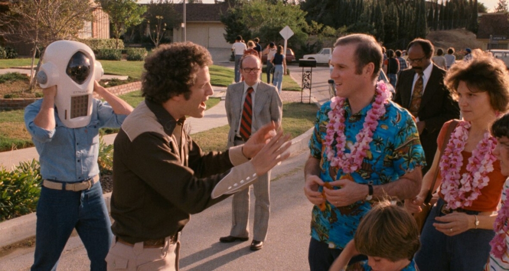 A man holding a peculiar helmet interacts with another man in a Hawaiian shirt and leis, surrounded by several people, including a child, on a suburban street.