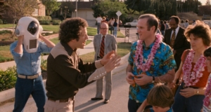 A man holding a peculiar helmet interacts with another man in a Hawaiian shirt and leis, surrounded by several people, including a child, on a suburban street.