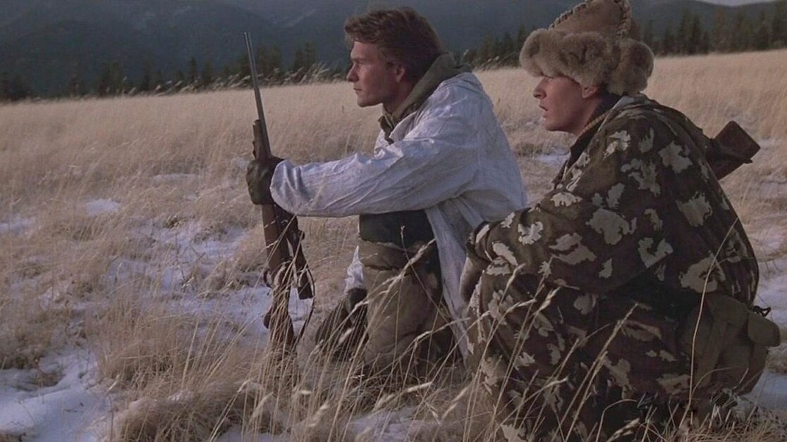 Two people, wearing camouflaged and winter gear, crouch in a snowy field with rifles, observing the landscape and mountains in the background.