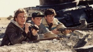 ‘Red Dawn’ (1984) 4K UHD Blu-Ray Review – Star-Studded Wartime Tale Is A Harrowing Experience Three young men crouch behind rocks, holding binoculars and rifles, looking intently into the distance.