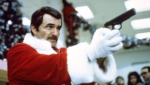 ‘Rent-A-Cop’ Blu-Ray Review – The Unlikely Team-Up Of Burt Reynolds And Liza Minnelli A man dressed as santa claus holding a gun with a focused expression in a crowded room.