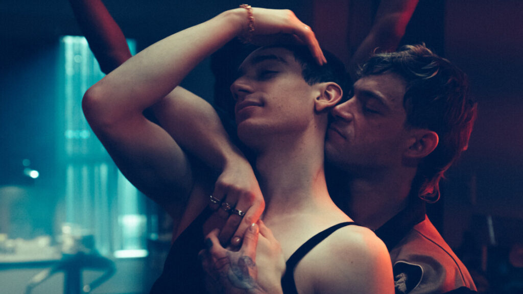 Two people embrace intimately in a dimly lit setting, with one person holding their partner from behind and both appearing content.
