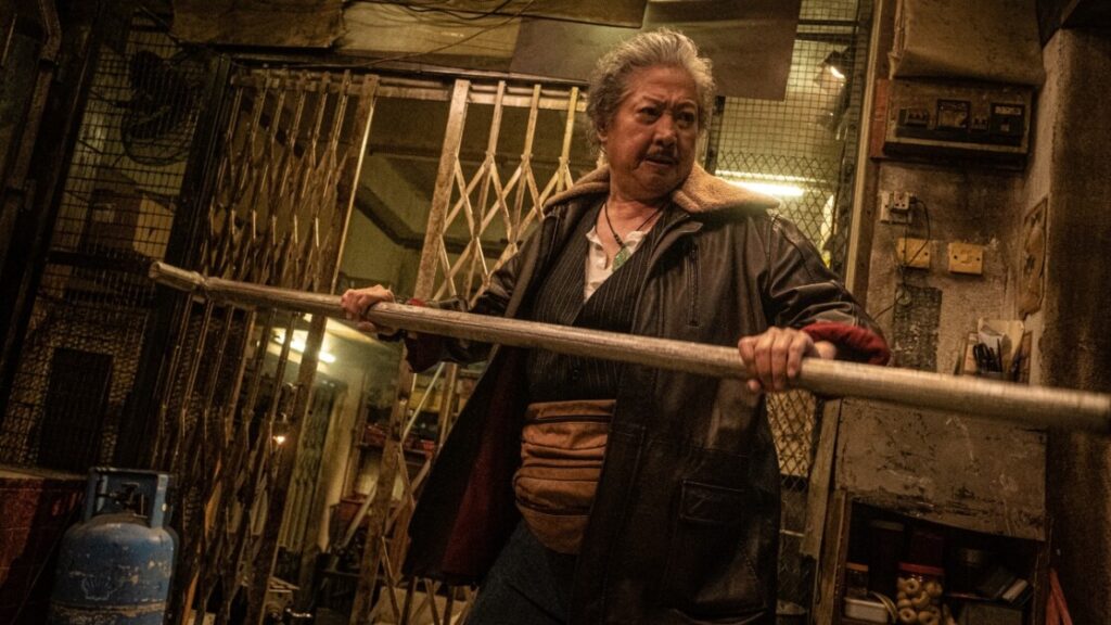 An older man with gray hair, wearing a leather jacket and fanny pack, holds a metal pipe in an industrial setting with metal gates and a blue gas cylinder.
