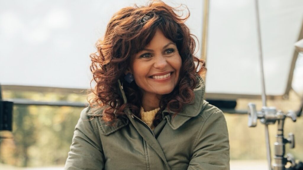 Woman with curly brown hair, wearing a green jacket, smiling outdoors with lighting equipment in the background.
