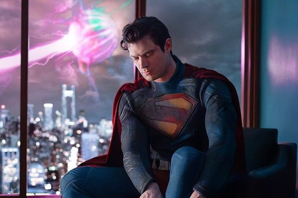 Superman in costume, sitting thoughtfully by a window with a neon-lit cityscape and a colorful spectral figure outside.