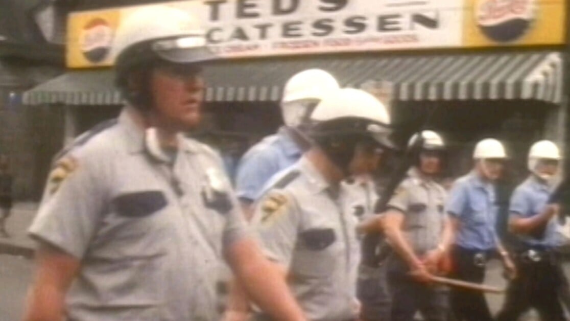 Police officers in helmets walking in front of Ted's Cateressen with batons, in an urban setting.
