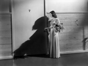A woman in a long dress holding a bouquet stands beside a wall, casting a large shadow. The number 7 is visible above her.