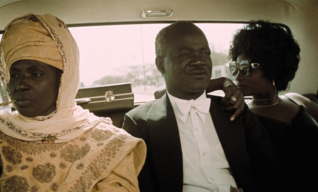 Three people are seated in the back of a car. The person on the left wears traditional attire, the person in the middle is in formal wear, and the person on the right is dressed in dark, modern clothing.