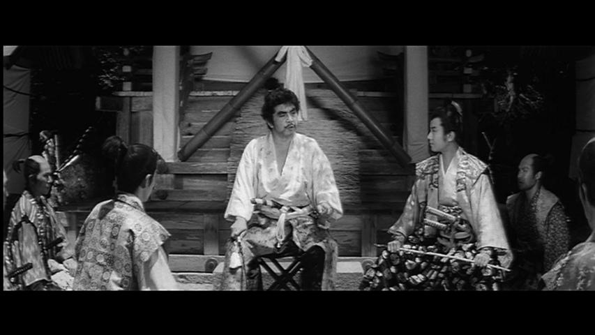 Black and white image from a film showing a man in traditional Japanese attire seated centrally, with several other characters in elaborate costumes surrounding him.