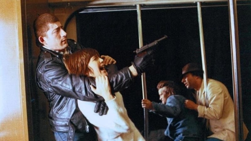 A scene depicts a man in a leather jacket holding a person in a white sweater at gunpoint on public transportation. Two other individuals in the background appear to be in a struggle.