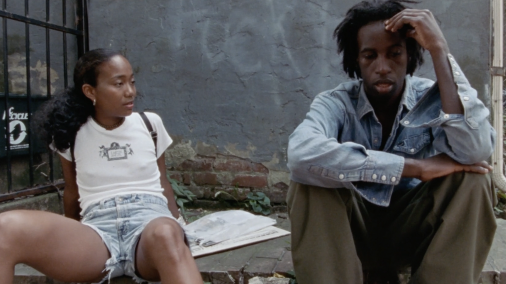 A young woman and man sit outside against a worn wall. The woman wears a white shirt and denim shorts, while the man wears a denim jacket and olive pants. They appear to be engaged in conversation.