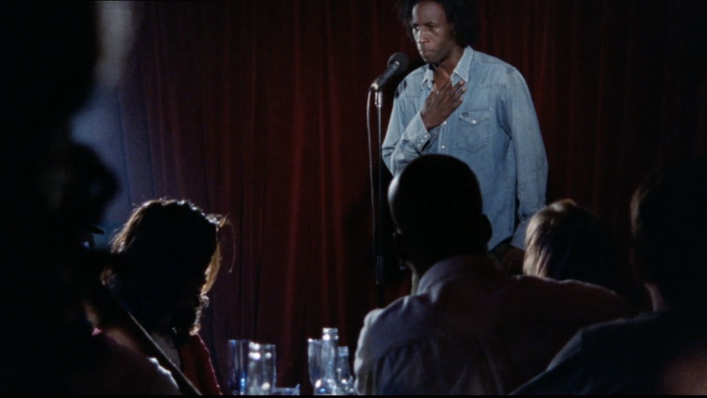 A person stands on stage speaking into a microphone, with an audience seated in front of them. The stage is backed by a red curtain.