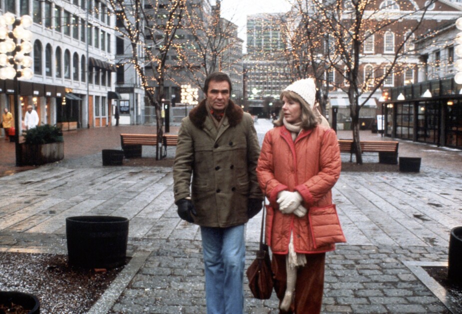 A man and woman walking through a city square lined with trees adorned with twinkling lights, wearing winter coats.