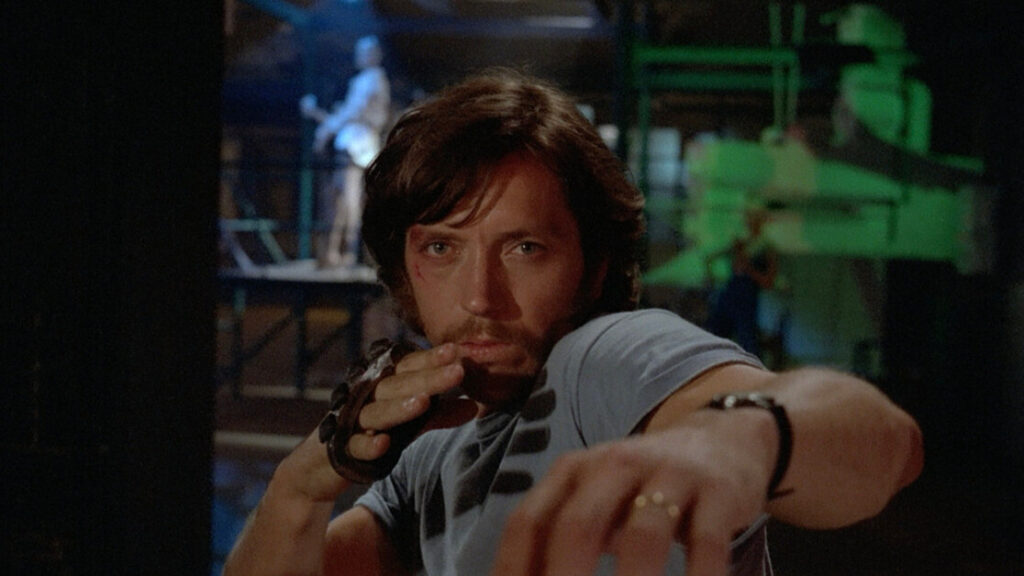 A man with long hair and a beard strikes a martial arts pose in a dimly lit setting. He wears a fingerless glove and appears focused, with another figure visible in the background.