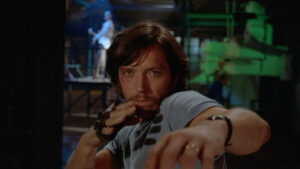Seeing Red: 3 French Vigilante Thrillers Blu-Ray Review – A Trio Of Distinct, Riveting Revenge Tales A man with long hair and a beard strikes a martial arts pose in a dimly lit setting. He wears a fingerless glove and appears focused, with another figure visible in the background.