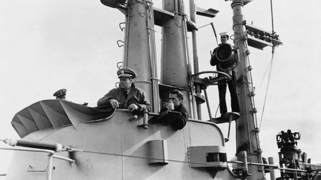 Two sailors in uniform aboard a naval ship, sitting in front of a large cylindrical structure with various naval equipment around them.