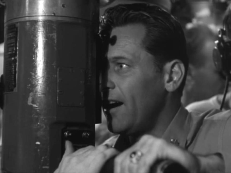 Black and white image of a man in a military uniform looking through a periscope with an intense expression.