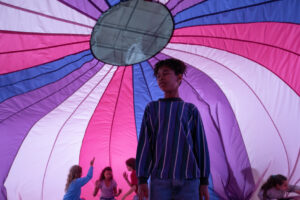 A group of children stand underneath a large, colorful parachute with one child in the foreground looking up.