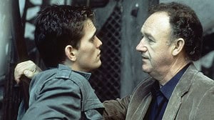 Two men having a serious conversation; a younger man with an intense expression holds an older man by the collar against a metal grate background.