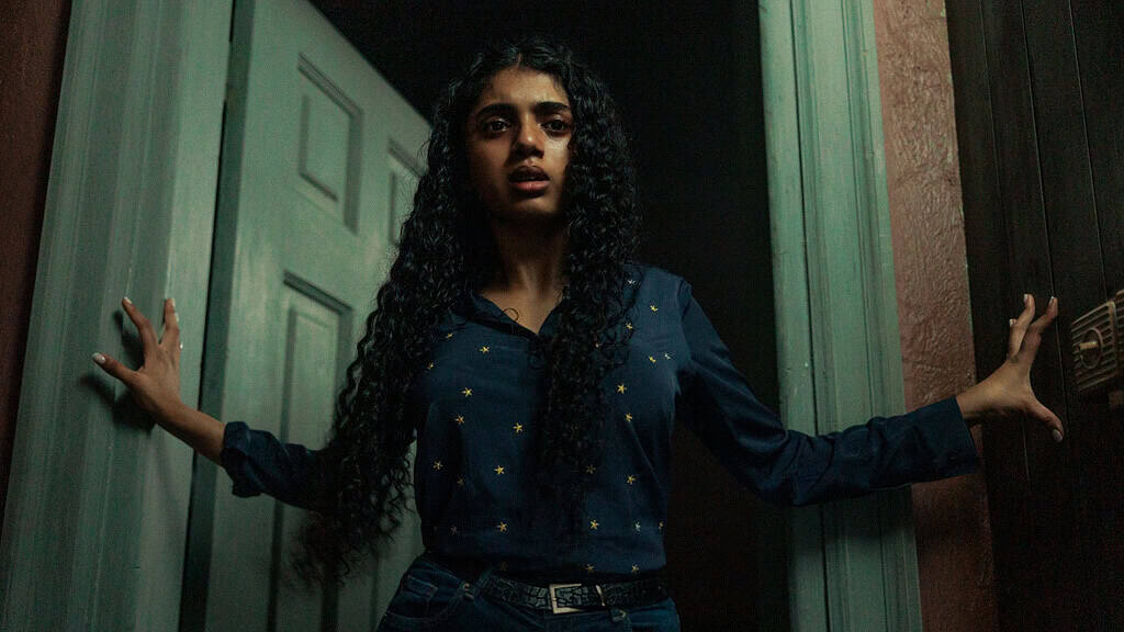 A woman with long curly hair stands apprehensively between two doors in dim lighting.