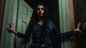 A woman with long curly hair stands apprehensively between two doors in dim lighting.