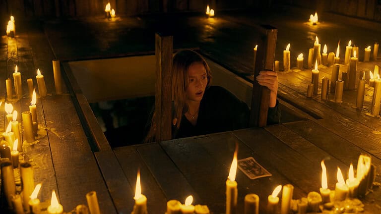 A woman emerges from a trapdoor at a candle-lit table, appearing anxious, surrounded by numerous lit candles in a dimly lit room.