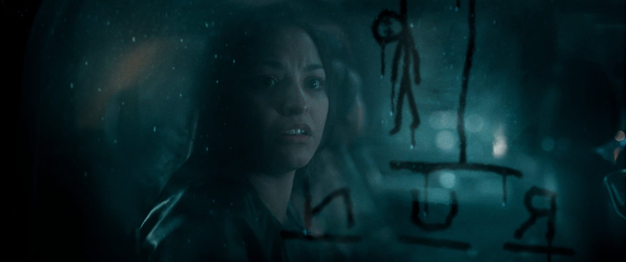 A woman looks anxious as she stares out a foggy window with ominous symbols and a stick figure drawn on it.