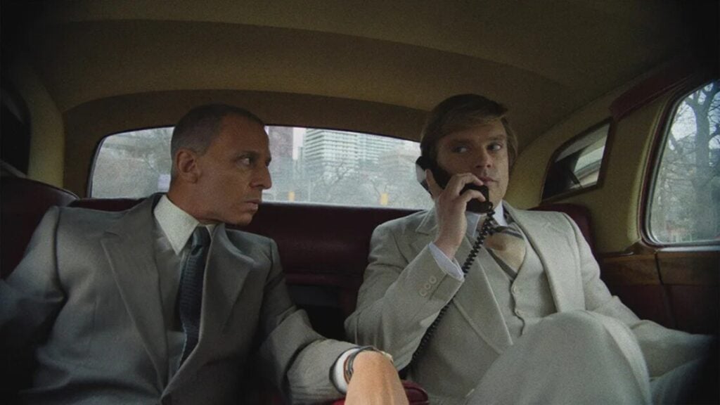 Two men in suits sit in the back of a vintage car. The man on the left looks at the man on the right, who is holding a phone to his ear.